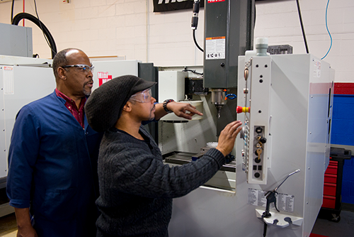 Monroe Community College student Geneo Brown operates a CNC machine as instructor Anthony McCollough watches